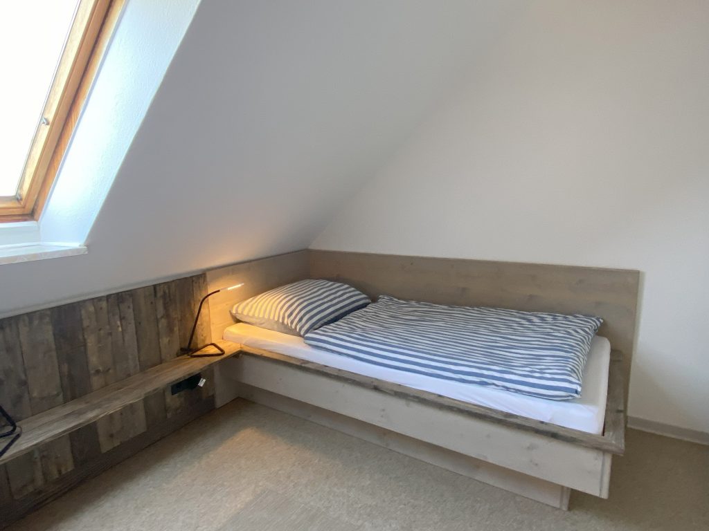 Blick ins Schlafzimmer auf eins der zwei handgefertigten Einzelbetten unter der Dachschräge. Licht fällt durch das Dachfenster.