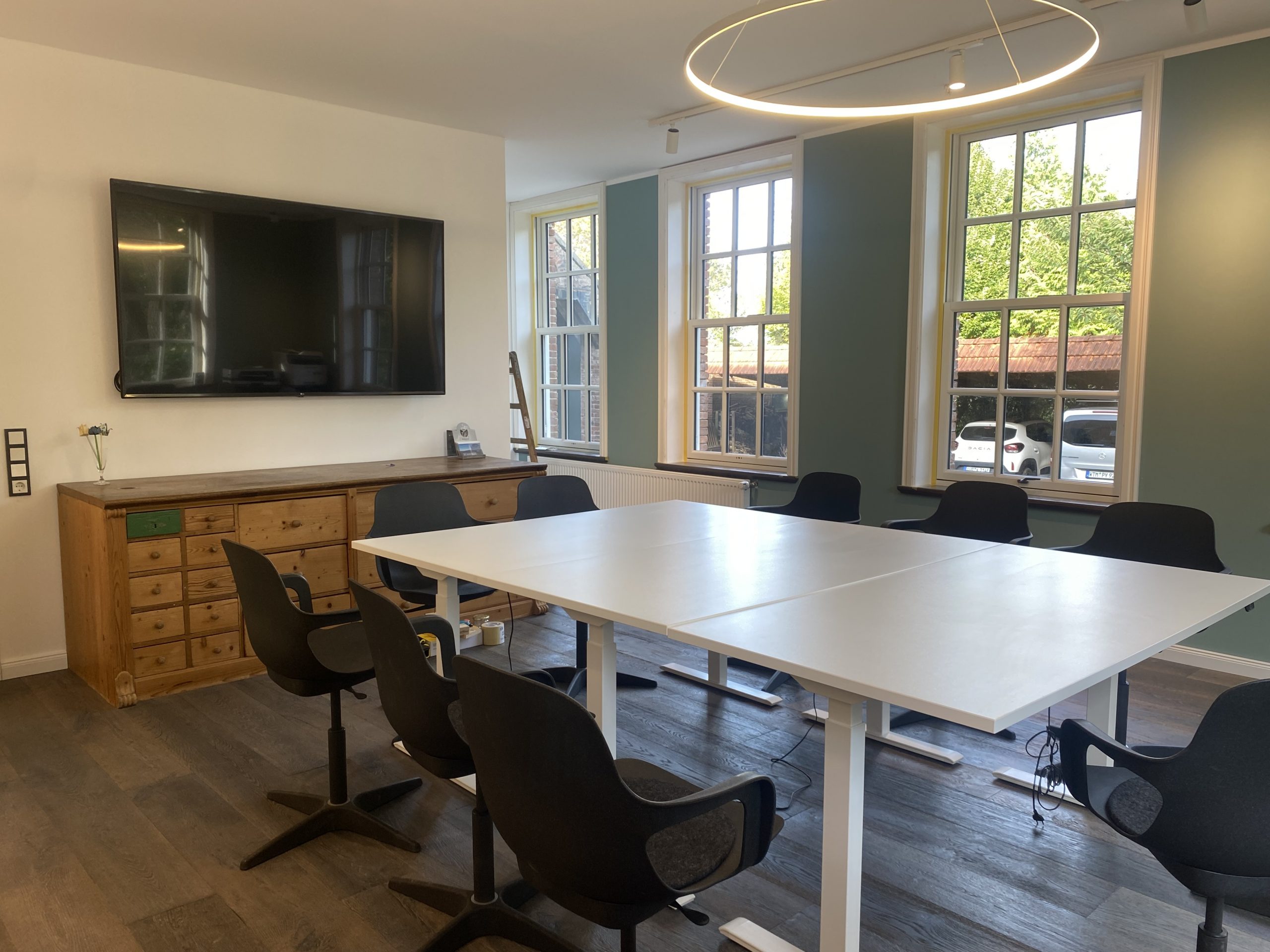 Im hellen Raum mit hohen Decken und großen Sprossenfenstern stehen mittig Tische und Stühle in Blockbestuhlung. Im Hintergrund ein großer Bildschirm an der Wand sowie eine große schwere Holzkommode.
