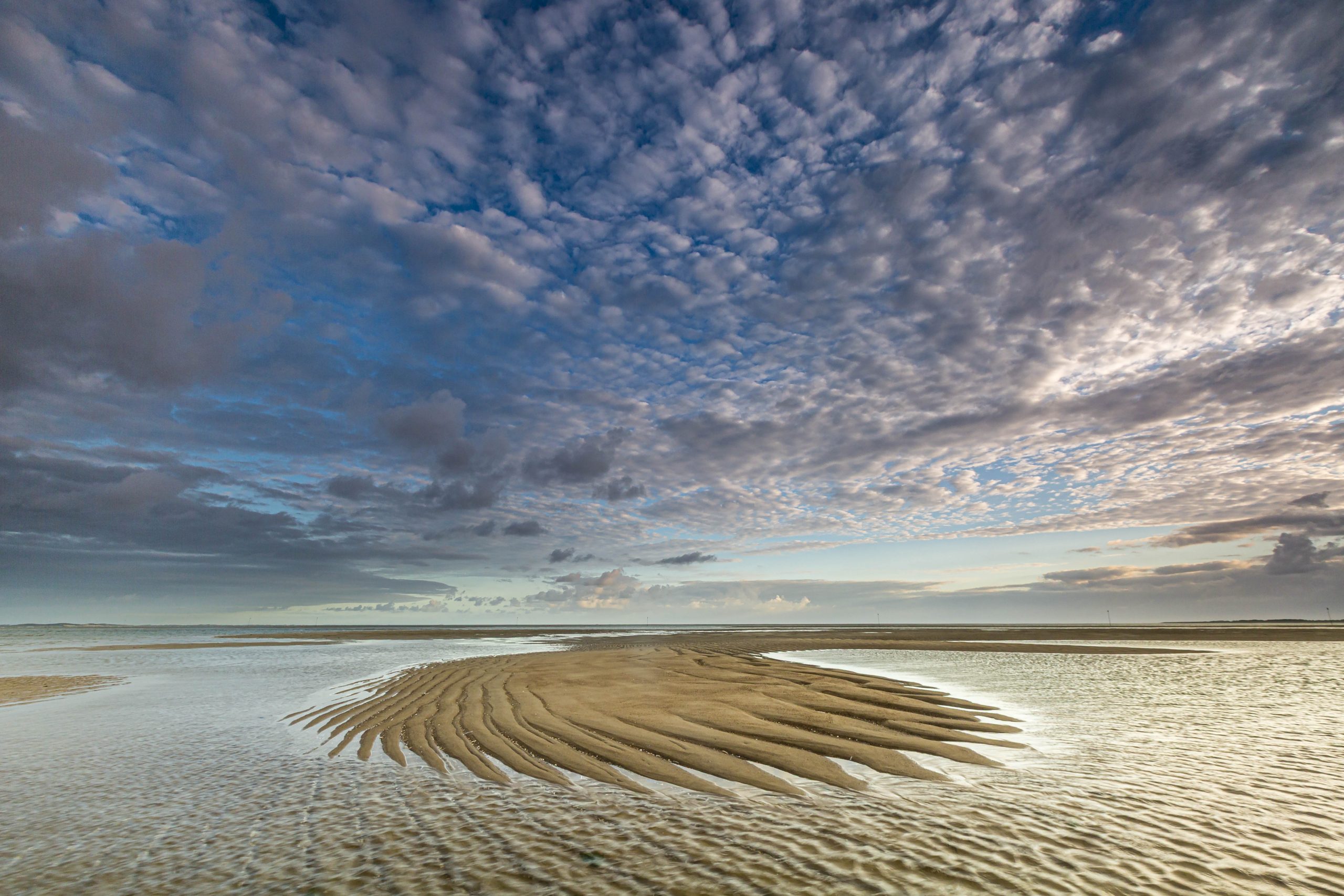 weite Wattlandschaft mit Wellenmuster am Boden und kleinen Wolken am blauen Himmel